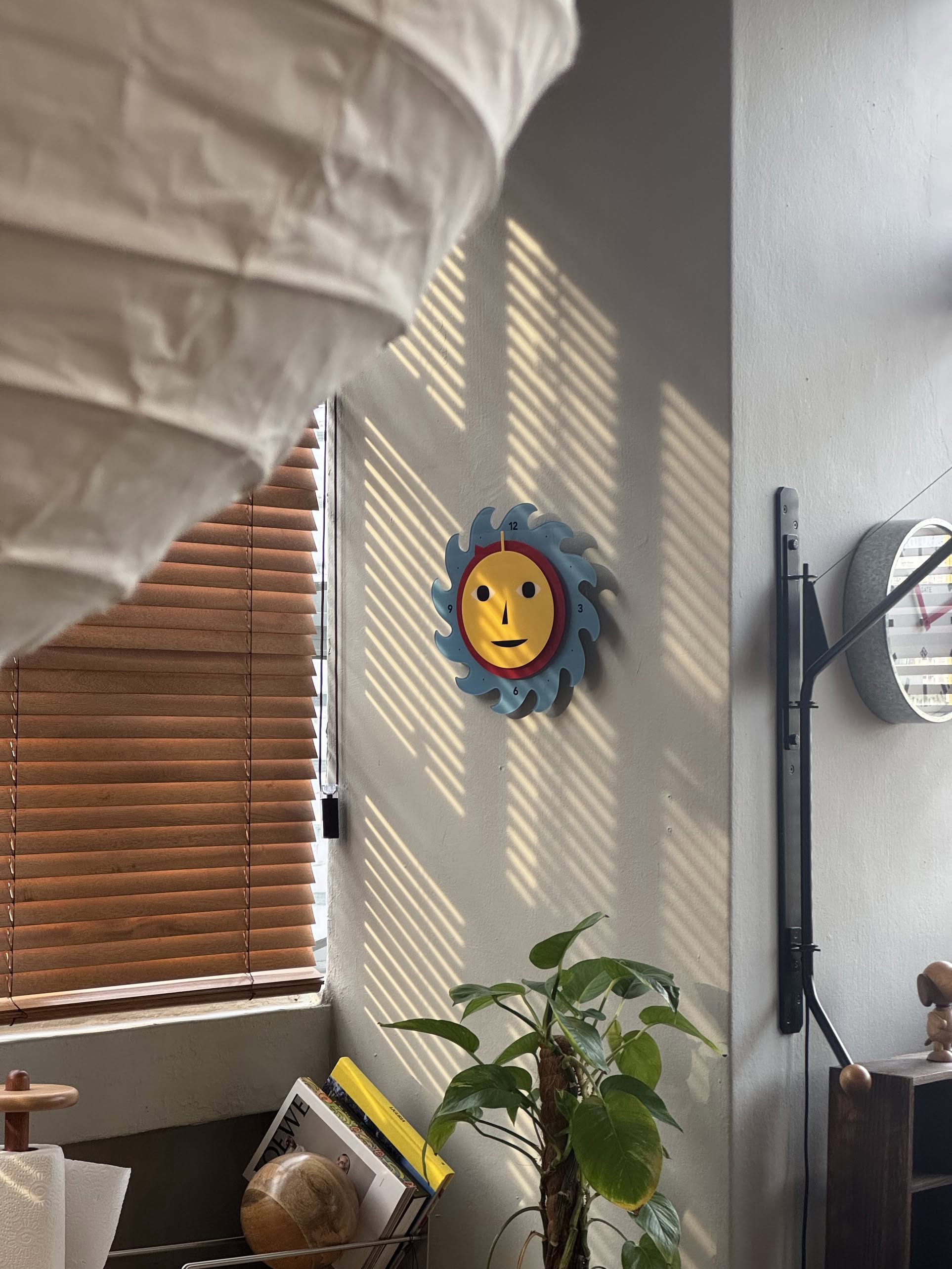 An Areaware Everybody Clock with a blue and yellow sun-shaped design by Areaware hangs on the wall, casting shadows from window blinds. Below, a green potted plant stands near books and a globe while a paper lantern brightens the foreground.