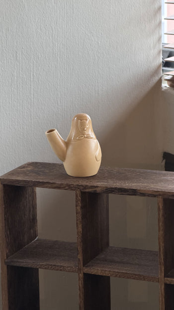 The Artek Easter Dog Vase - Sand by Artek, shaped like a person, sits on a wooden shelf near a window with blinds—a subtle nod to Finnish Easter tradition against a light-colored wall.