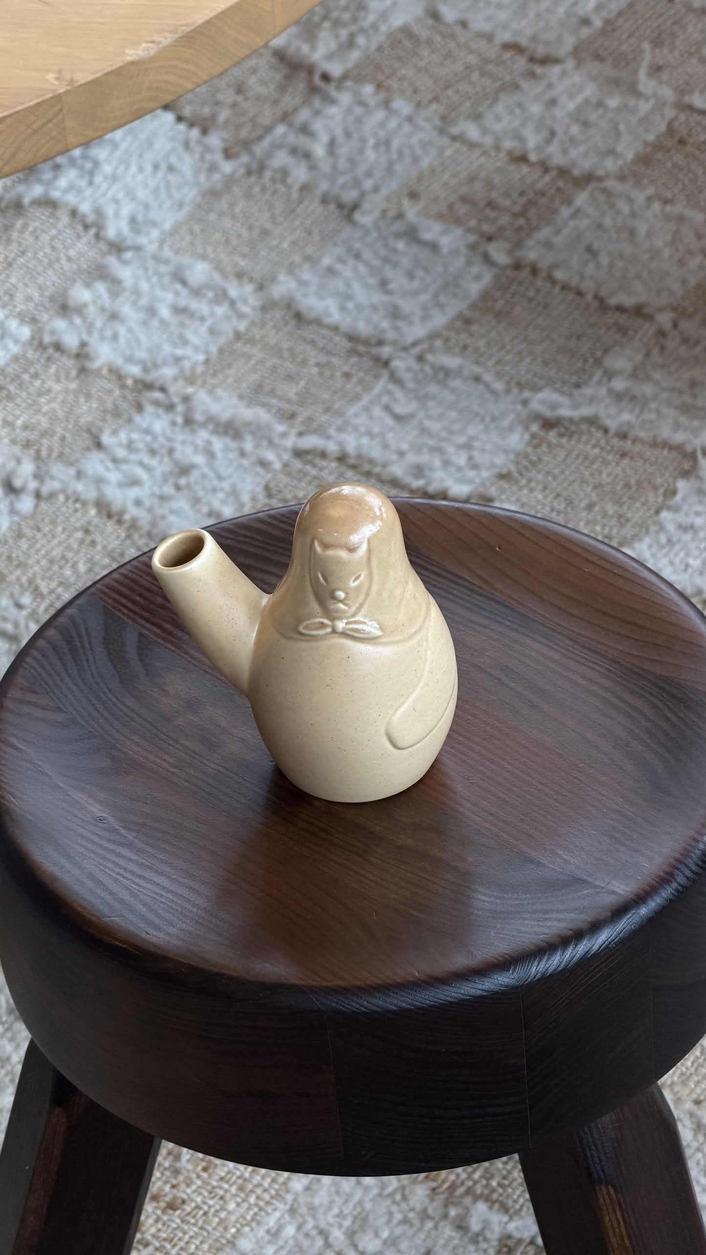 A sand-toned Artek Easter Dog Vase by Artek, reminiscent of Finnish Easter traditions, sits on a dark wooden stool with a textured, patterned rug in the background.