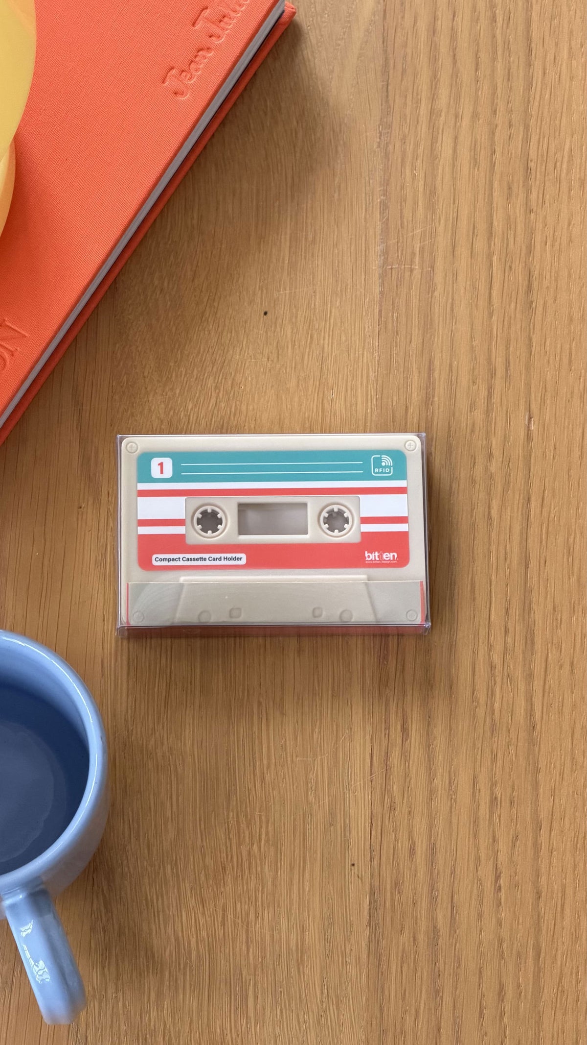 The Bitten Cassette Card Holder by Bitten rests on a wooden table beside a blue cup and a red hardcover book.