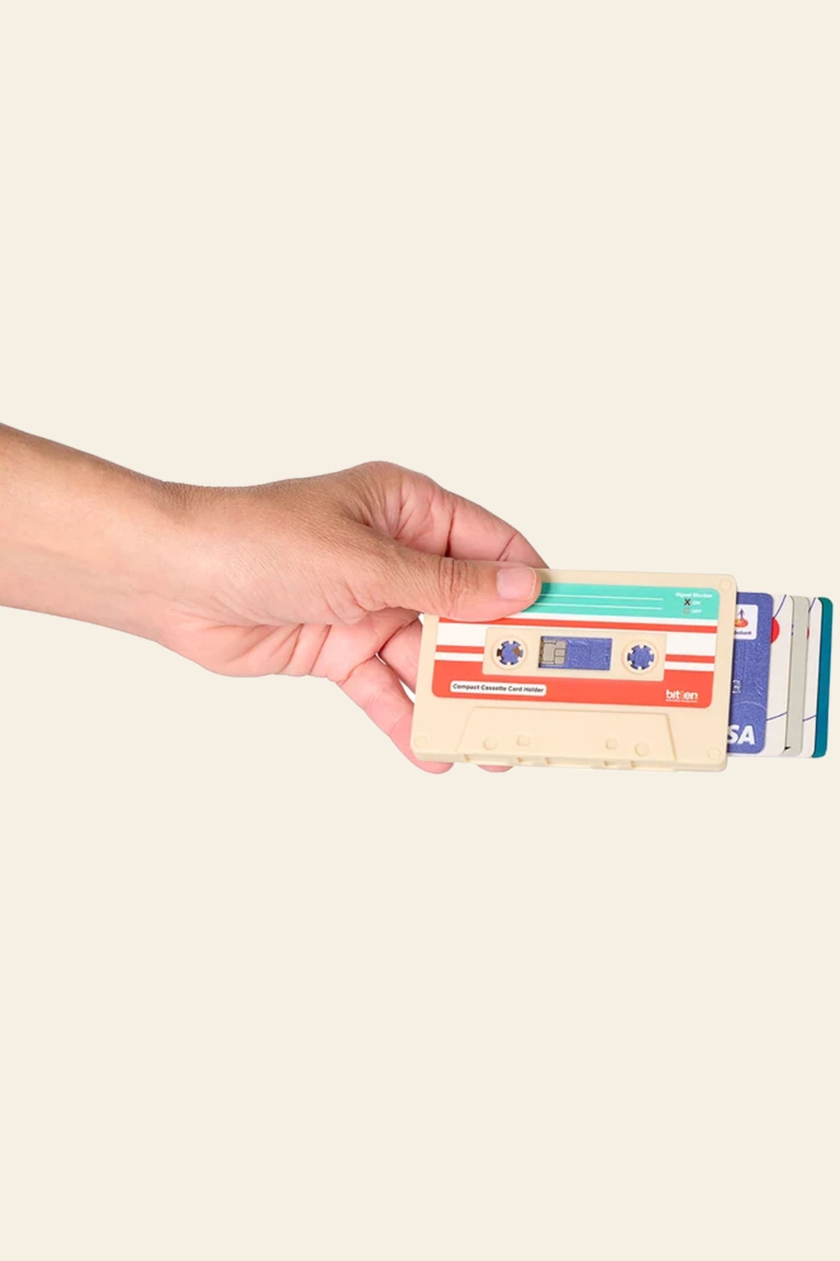 A hand holds the Bitten Cassette Card Holder by Bitten, with several cards—including a visible blue credit card—partially pulled out from the RFID holder against a beige background.