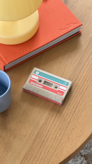 A Bitten Cassette Card Holder in a clear case sits on a wooden table near a blue cup, an orange book, and a yellow lamp.