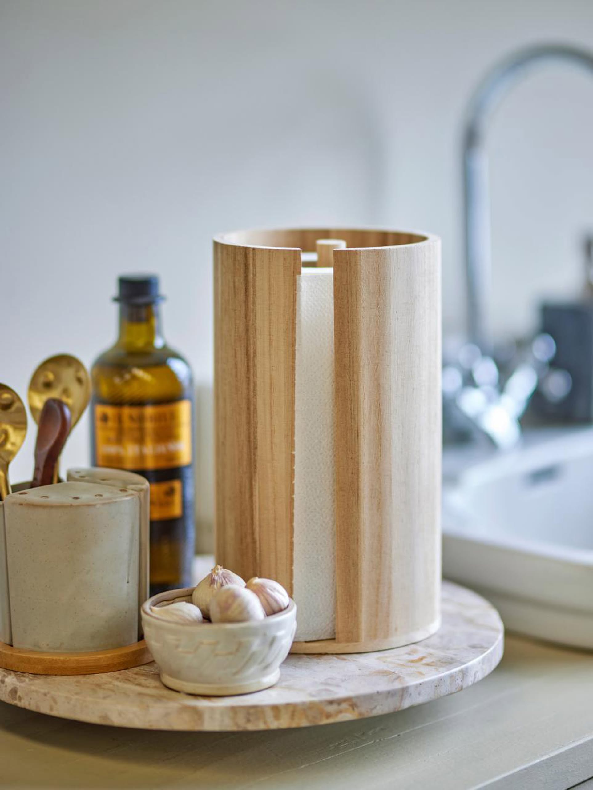 The Bloomingville Celian Kitchen Paper Stand sits on a round marble tray with a bowl of garlic, an oil bottle, and a utensil holder on a kitchen countertop near the sink.