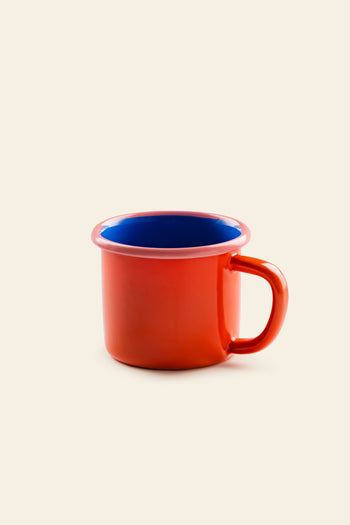 The Bornn Enamelware Colorama Mug by Bornn Enamelware features a vibrant red exterior and a deep blue interior. Its handle design sits elegantly against a plain light beige background, highlighting the mug’s bold and colorful contrast.
