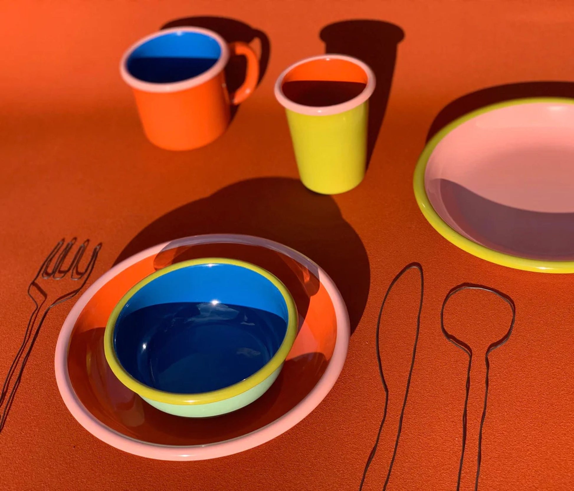 Bornn Enamelware Colorama Mugs from Bornn Enamelware are shown on an orange surface beside black-outlined fork, knife, and spoon drawings, creating a playful table setting ideal for coffee or casual meals.