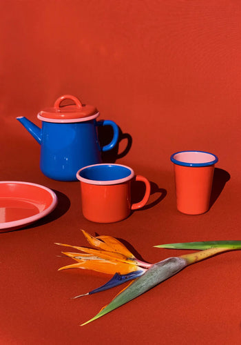 A Bornn Enamelware Colorama Mug sits on a red surface beside other vibrant enamelware and a bird of paradise flower—ideal for serving coffee or tea in bold, colorful style.