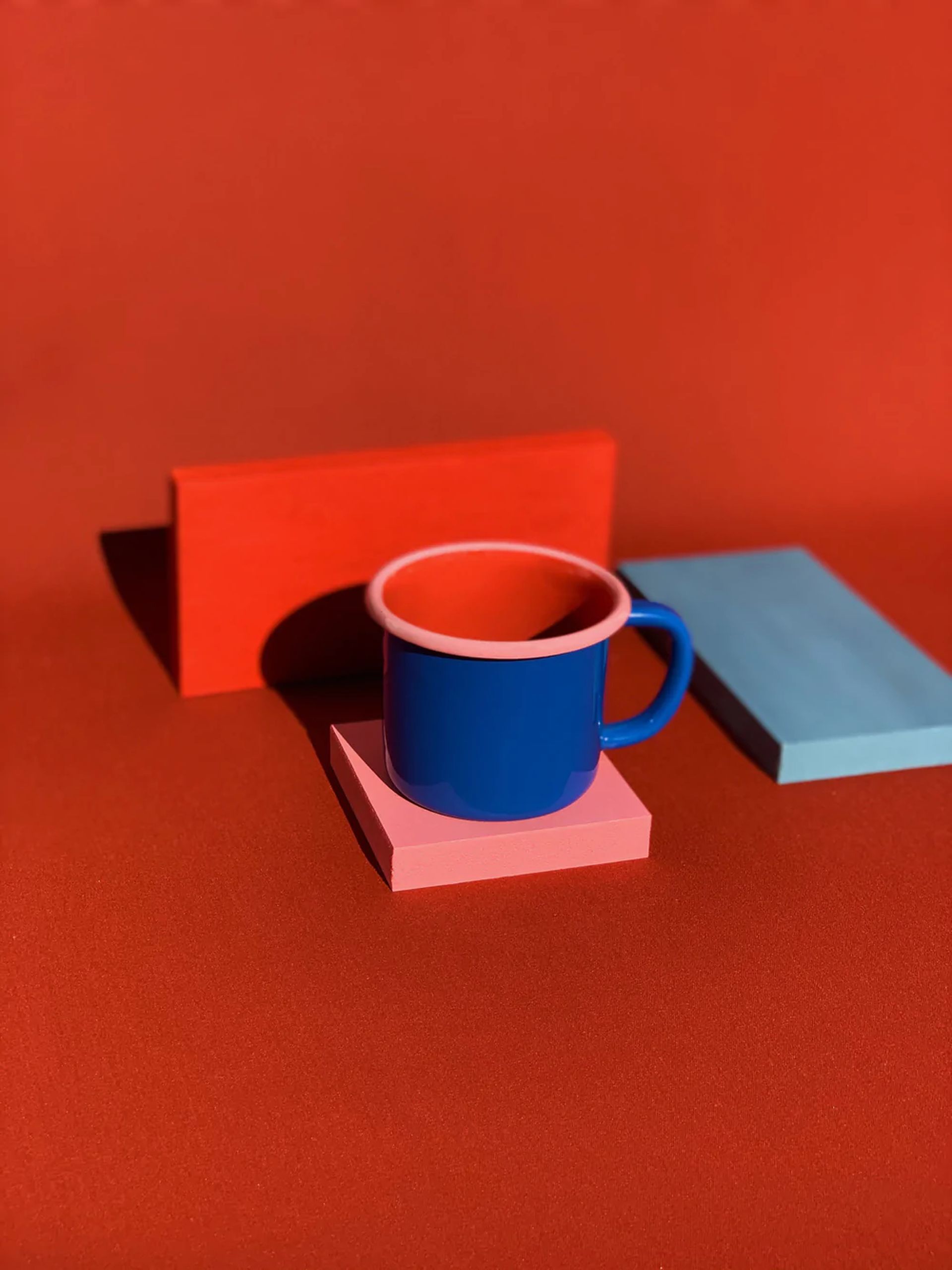 The Bornn Enamelware Colorama Mug by Bornn Enamelware features blue enamel with a pink rim, displayed on a pink block beside blue and red rectangles, set against a bold red background—ideal for coffee lovers.