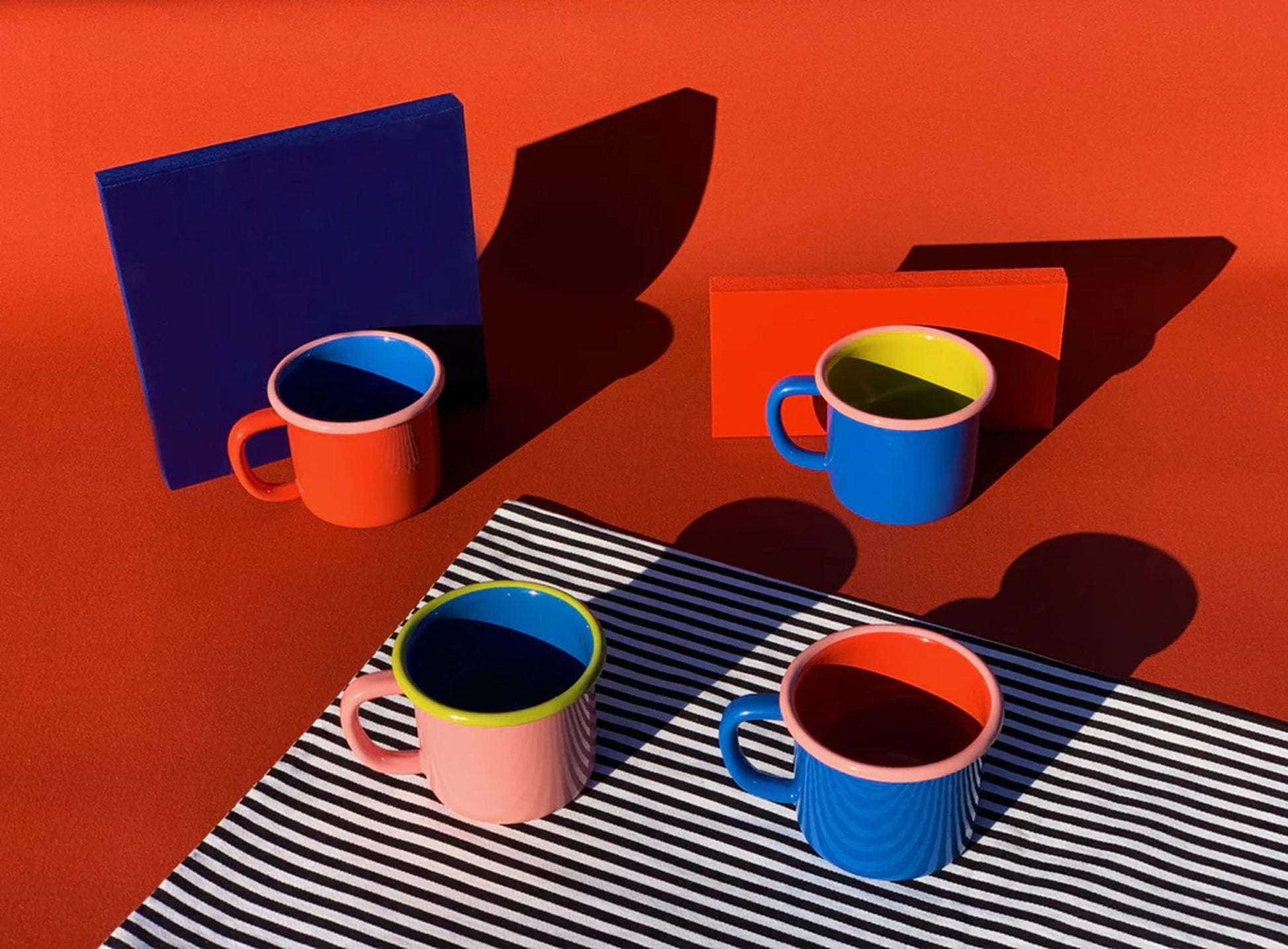 Four Bornn Enamelware Colorama Mugs in red, blue, pink, and blue-red sit on a red surface with two blocks and a striped cloth, casting bold shadows in sunlight. Brighten your next coffee break with Bornn Enamelware style!.