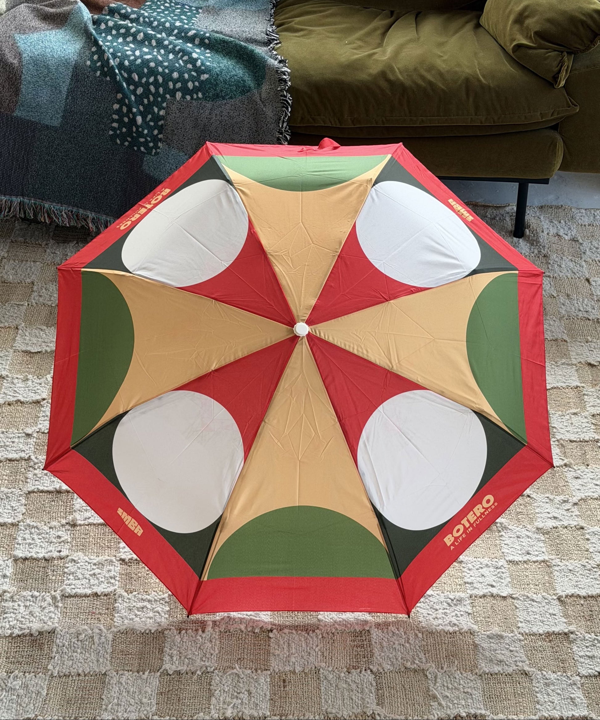 The Botero Patterned Umbrella in Red, Green, and Yellow by Botero is open on a beige patterned rug beside a green sofa and a fringed, multicolored blanket.