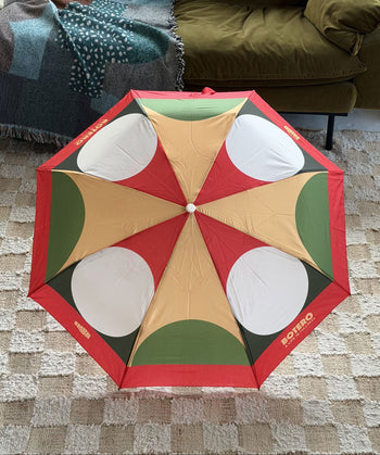 The Botero Patterned Umbrella in Red, Green, and Yellow by Botero is open on a beige patterned rug beside a green sofa and a fringed, multicolored blanket.