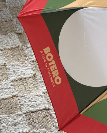 A vibrant Botero Patterned Umbrella in red, green, and yellow—featuring the text “BOTERO A LIFE IN FULLNESS” on a red panel—rests on a textured woven rug. This eye-catching accessory from Botero is both stylish and practical, perfect for brightening up rainy days.
