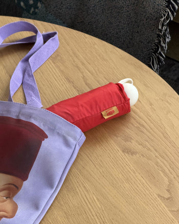A Botero Patterned Umbrella in red, partially sticking out of a light purple tote bag with a printed image, rests on a wooden table.
