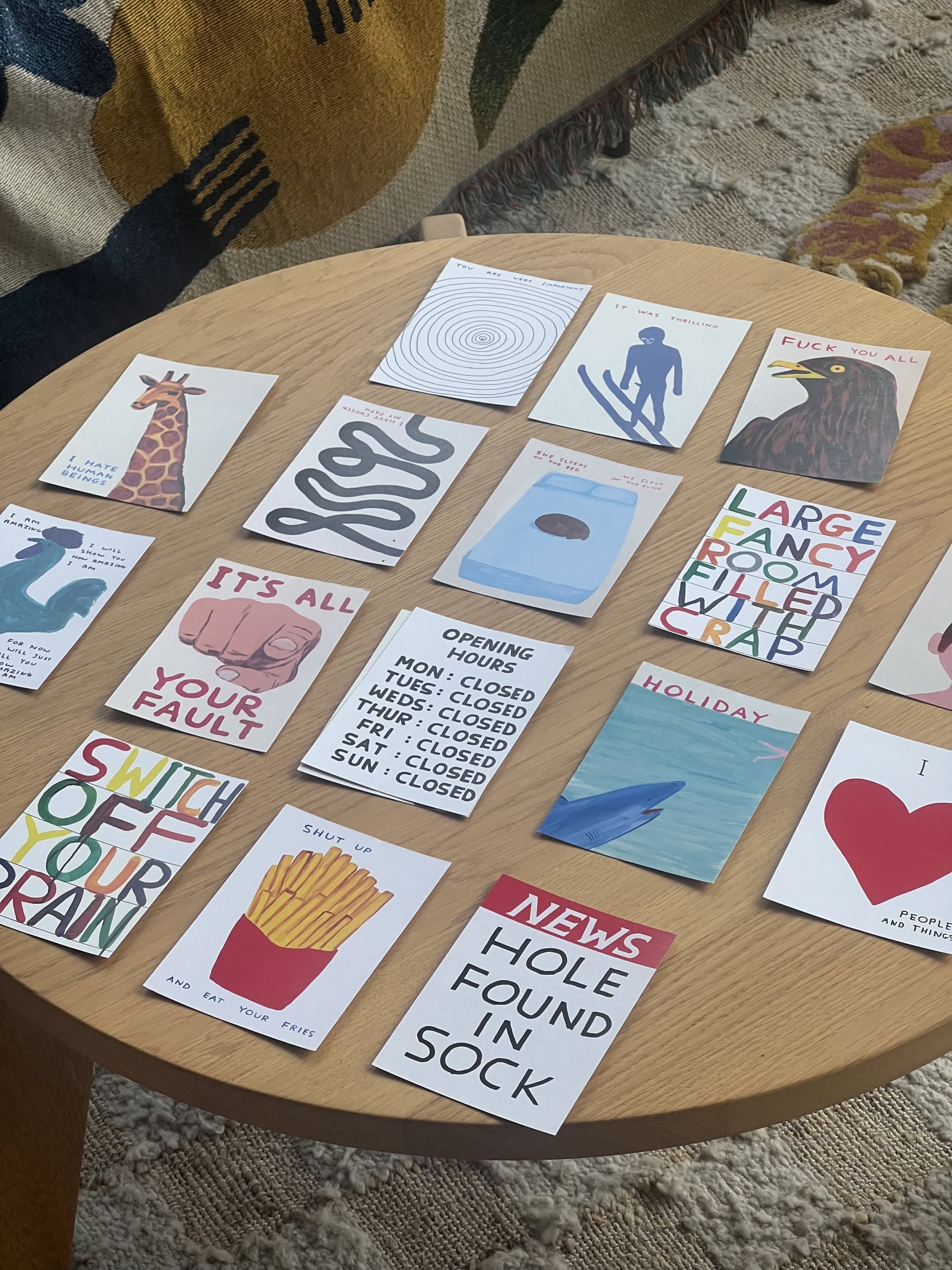 A round wooden table displays 15 illustrated greeting cards with humorous, playful designs—ideal for holiday gifting or as a personalized option like the Stacked Store Brainbox Candy Postcard Gift Card.