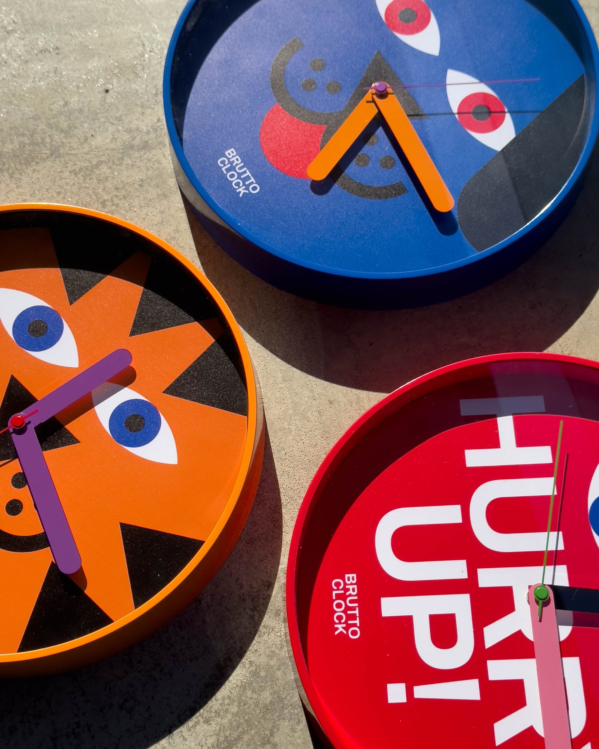 Three Brutto Studio multicolour wall clocks with playful designs lie on concrete: one Hurry Up! Wall Clock and two with dog and cat faces. Each features pink and orange hands and silent quartz movement.