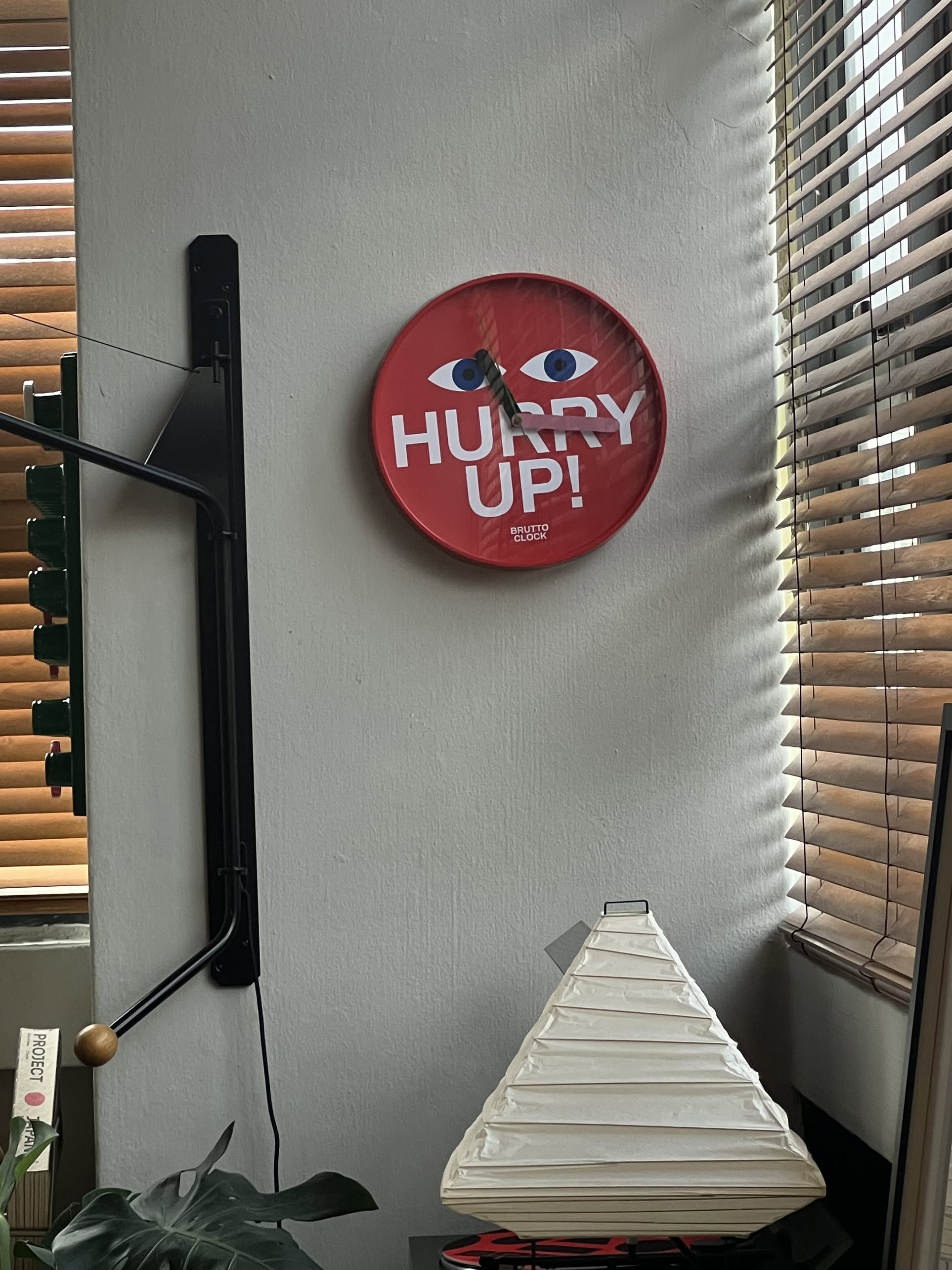 The Brutto Studio Hurry Up! Wall Clock - Multicolour, with googly eyes and bold letters, hangs on a white wall near two blinds-covered windows. Its silent quartz movement ensures quietness; below is a white lampshade and a plant peeks from the left.