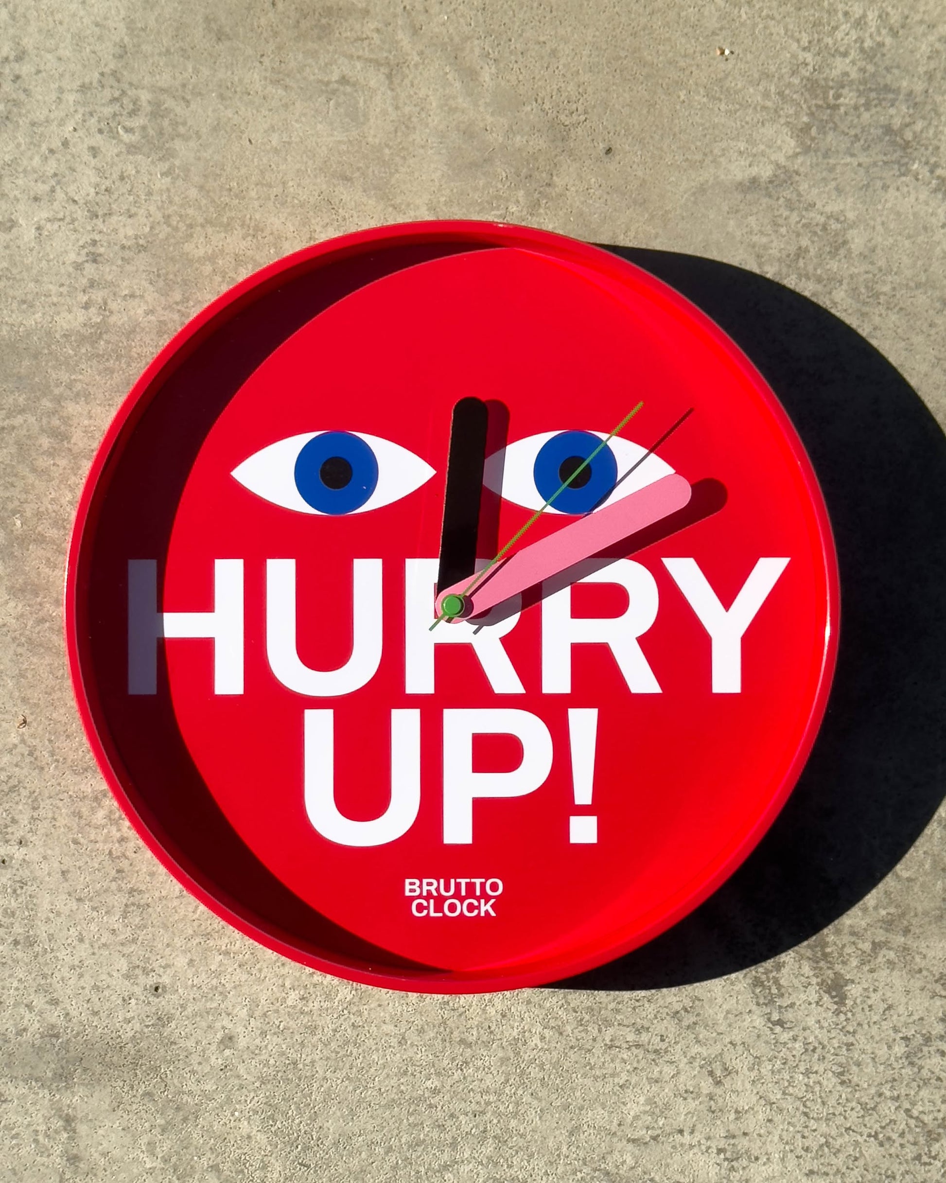 The Brutto Studio Hurry Up! Wall Clock – Multicolour features stylized eyes above bold white "HURRY UP!" text and "BRUTTO CLOCK" below. Silent quartz movement ensures quiet operation. Clock hands are set to 2:00 on a concrete surface.