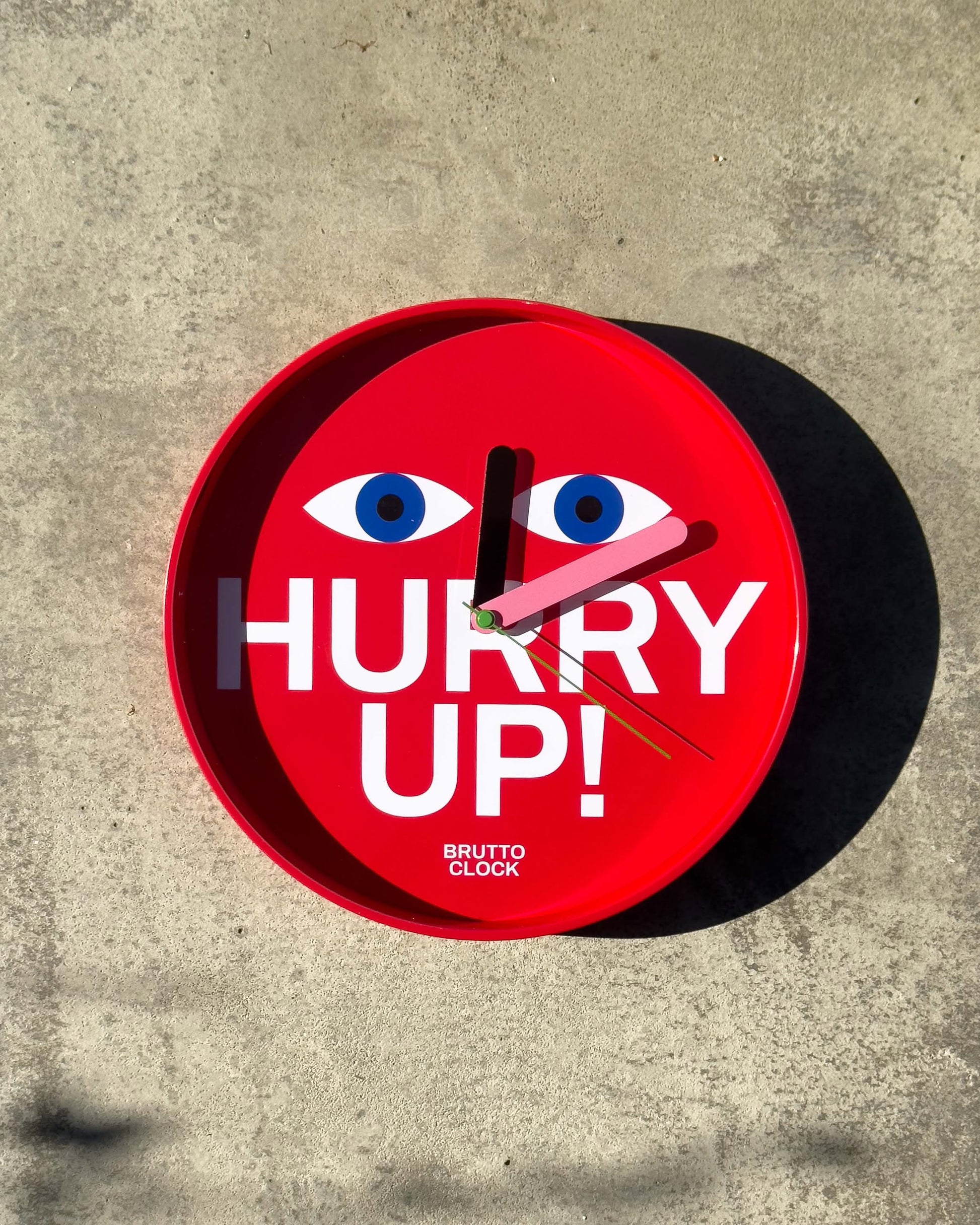 The Brutto Studio Hurry Up! Wall Clock - Multicolour by Brutto Studio features bold white letters, blue cartoon eyes, “BRUTTO CLOCK” at the bottom, shows 10:09, casts a shadow on concrete, and has a silent quartz movement for precision.