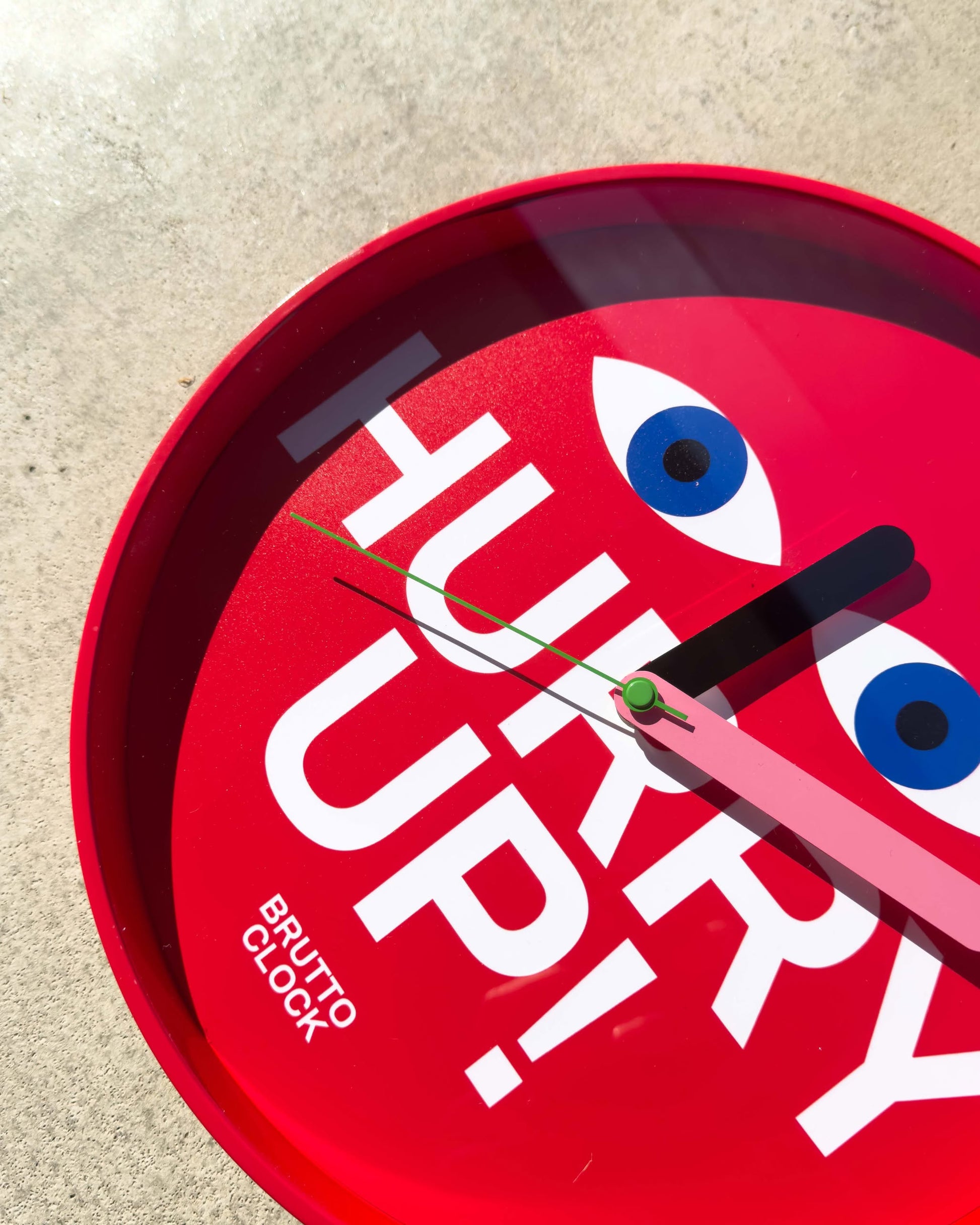 The Brutto Studio Hurry Up! Wall Clock - Multicolour by Brutto Studio, with cartoon eyes and bold “HURRY UP!” text, is shown against a light concrete background.