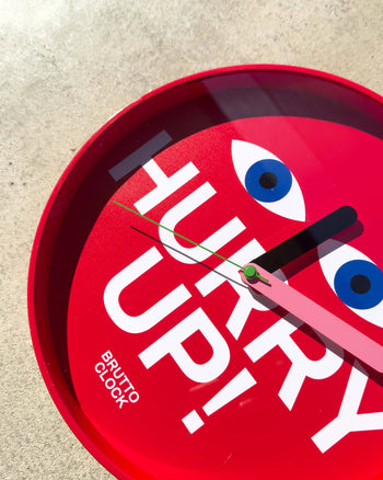 The Brutto Studio Hurry Up! Wall Clock - Multicolour by Brutto Studio, with cartoon eyes and bold “HURRY UP!” text, is shown against a light concrete background.