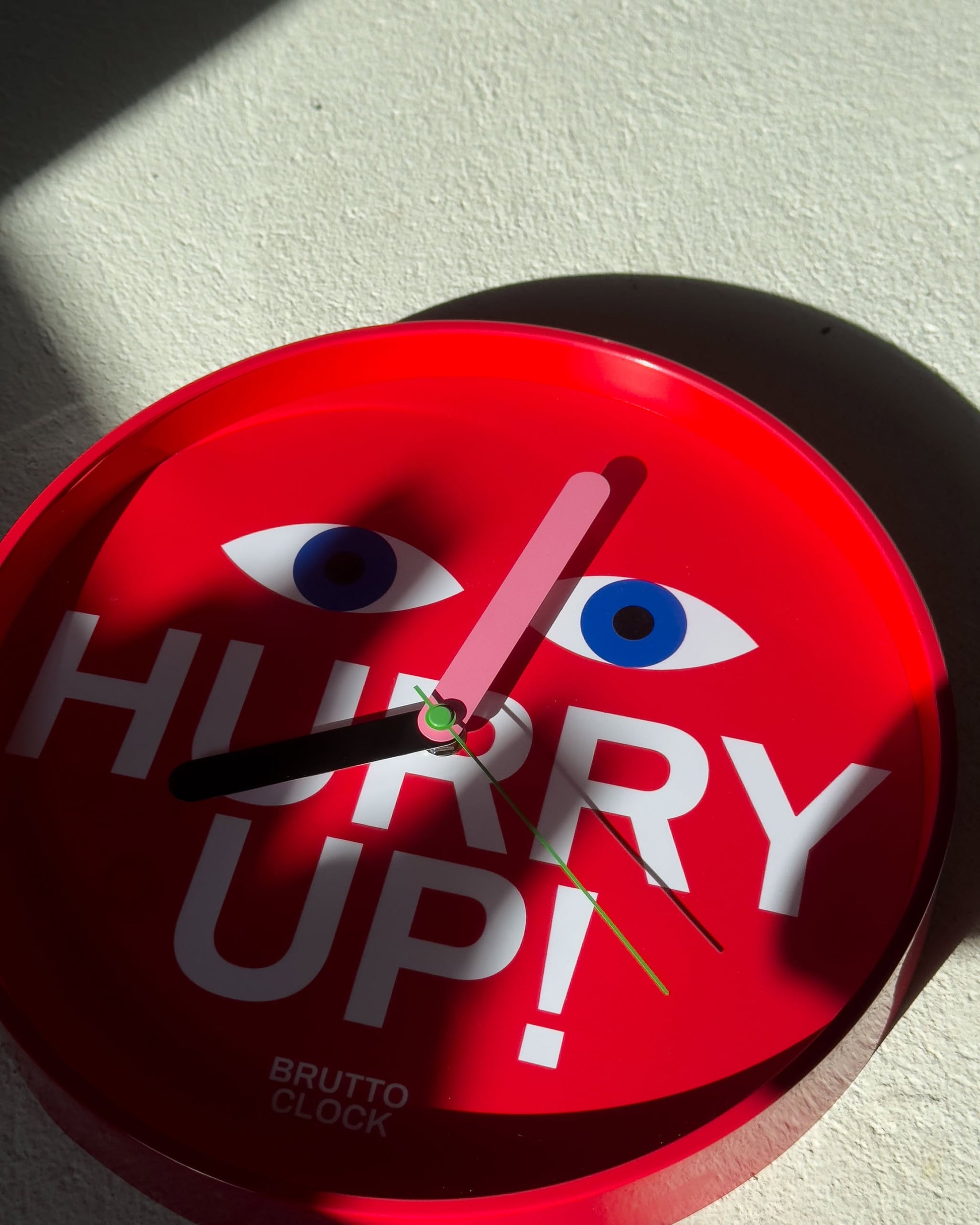 The Brutto Studio Hurry Up! Wall Clock - Multicolour features playful illustrated eyes above "HURRY UP!", silent quartz movement, and bright colours, shown at 10:05 with sunlight and shadow on a light surface.