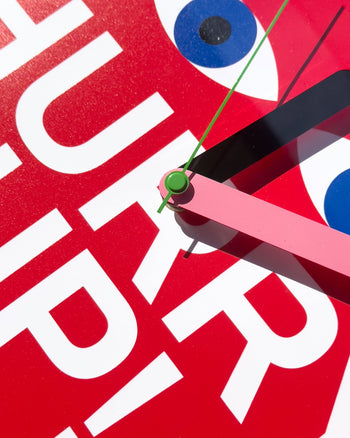 Close-up of the Brutto Studio Hurry Up! Wall Clock – Multicolour, featuring a bold red face, white text, blue and white eye-like circles, and pink and green hands for a playful touch in any room.