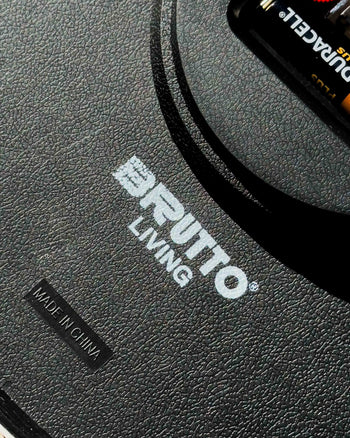 Close-up of a black textured surface with the white Brutto Studio logo, a MADE IN CHINA label, and part of a Duracell battery—perfect for your Brutto Studio Hurry Up! Wall Clock - Multicolour featuring silent quartz movement.