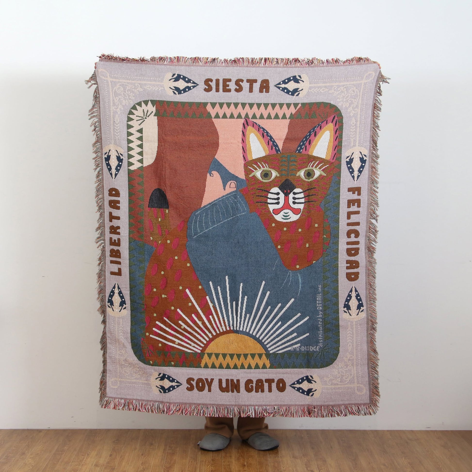 A person holds up the DETAIL Cat Oax Throwket by DETAIL, a vibrant throw blanket featuring colorful folk art cat motifs, Spanish words like “Siesta” and “Libertad,” and abstract shapes—only their feet are visible behind it.