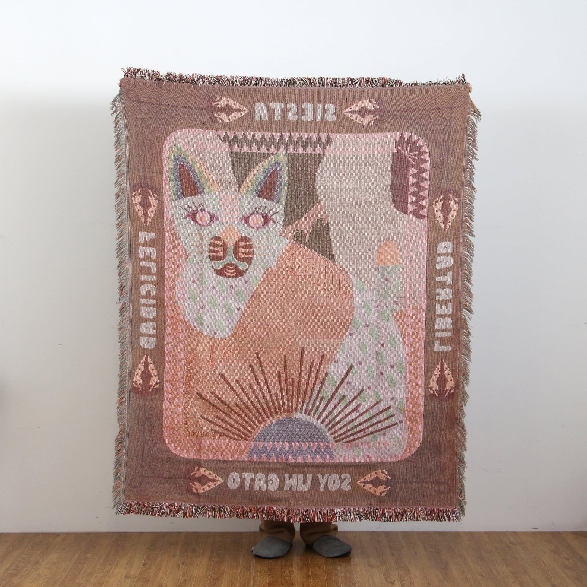 A person holds up the DETAIL Cat Oax Throwket by DETAIL, featuring a stylized cat, geometric shapes, SOY UN GATO text, and mirrored or rotated words, standing against a plain wall and wooden floor.
