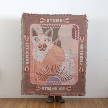 A person holds up the DETAIL Cat Oax Throwket by DETAIL, featuring a stylized cat, geometric shapes, SOY UN GATO text, and mirrored or rotated words, standing against a plain wall and wooden floor.