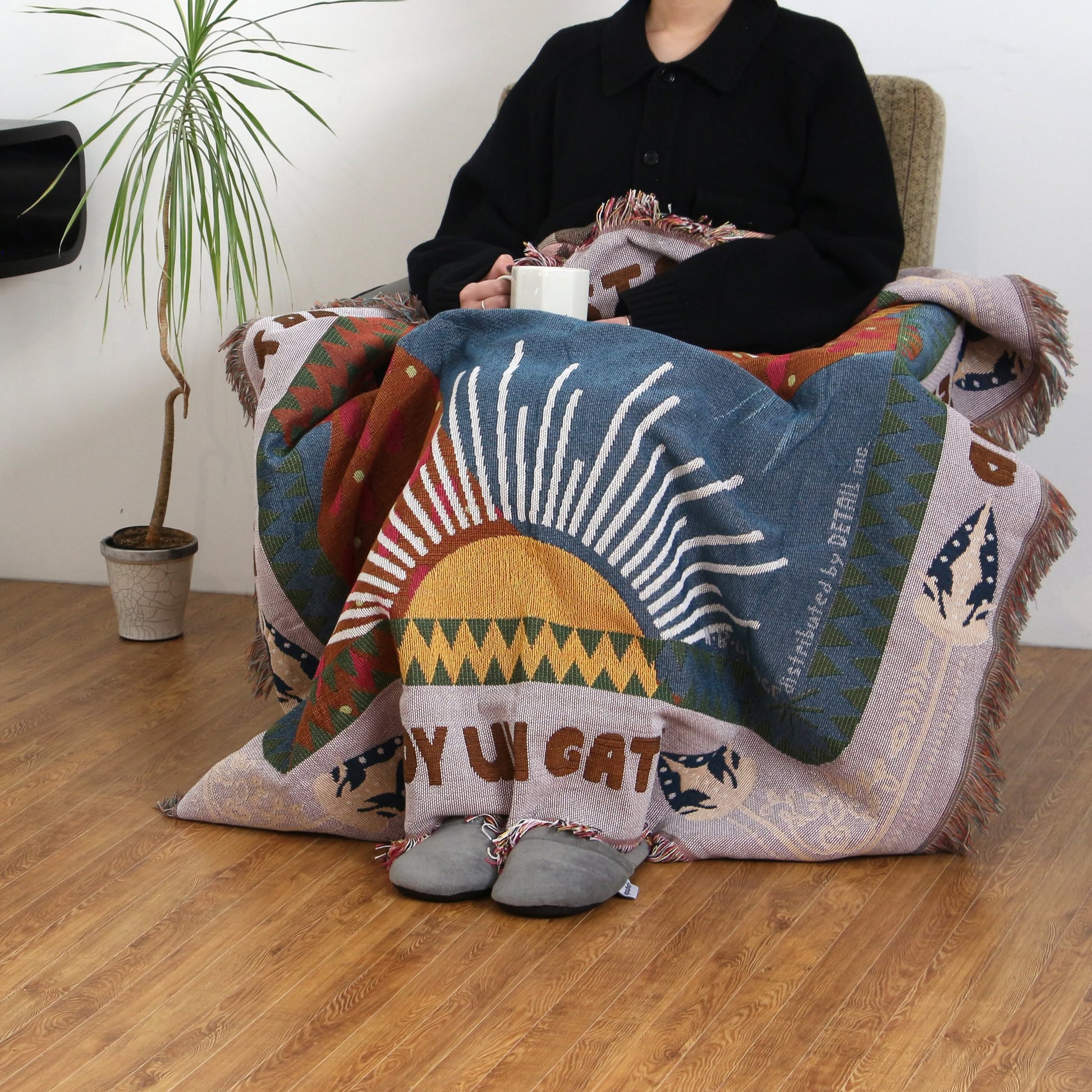 A person sits on a chair holding a white mug, wrapped in the DETAIL Cat Oax Throwket by DETAIL featuring sun and cat designs. The wooden floor and a nearby potted plant complete the scene, with their face not shown.