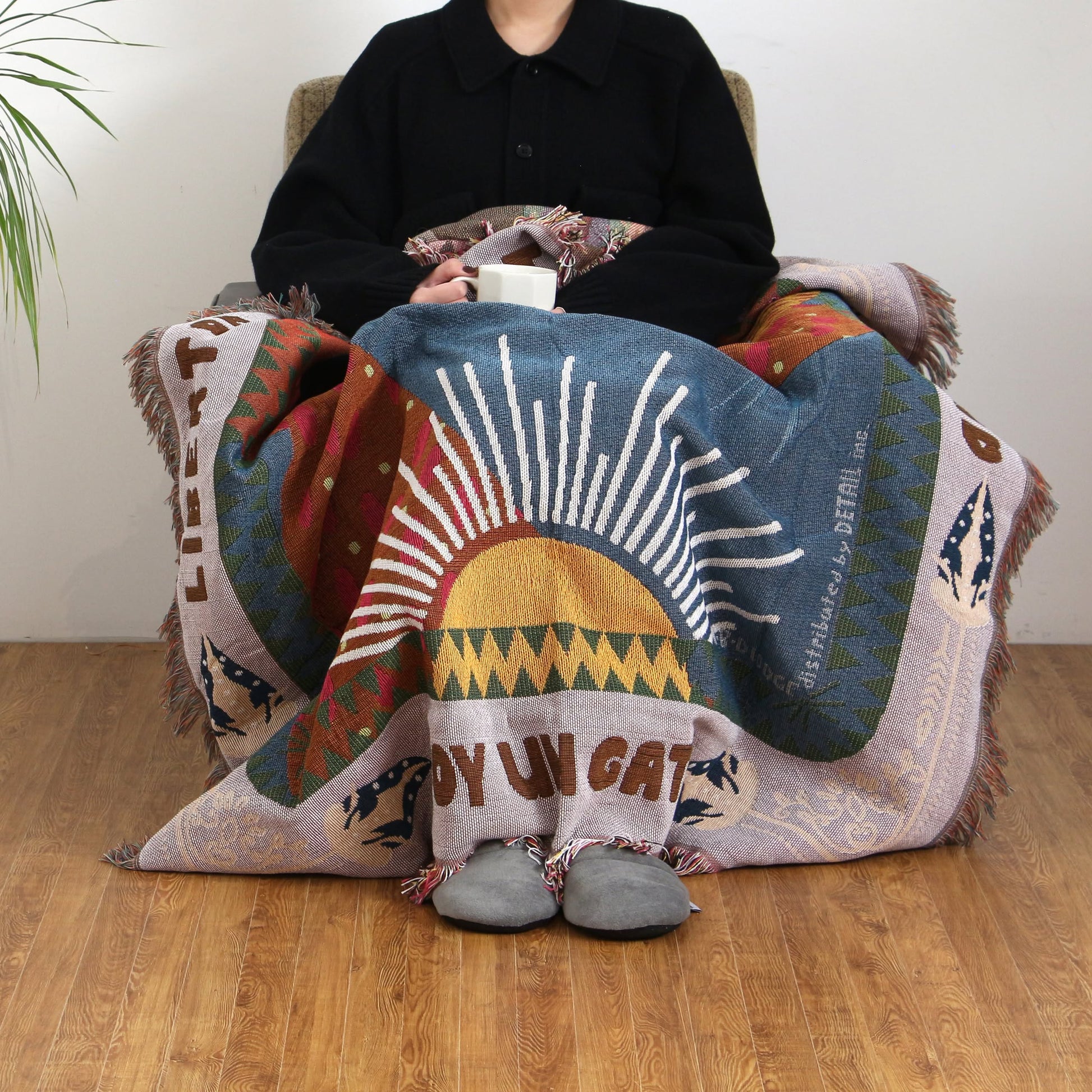 A person in a black coat sits on a chair with the DETAIL Cat Oax Throwket by DETAIL, featuring a sun design, draped over their lap while holding a white mug. The floor is wooden and theres a plant visible on the left.