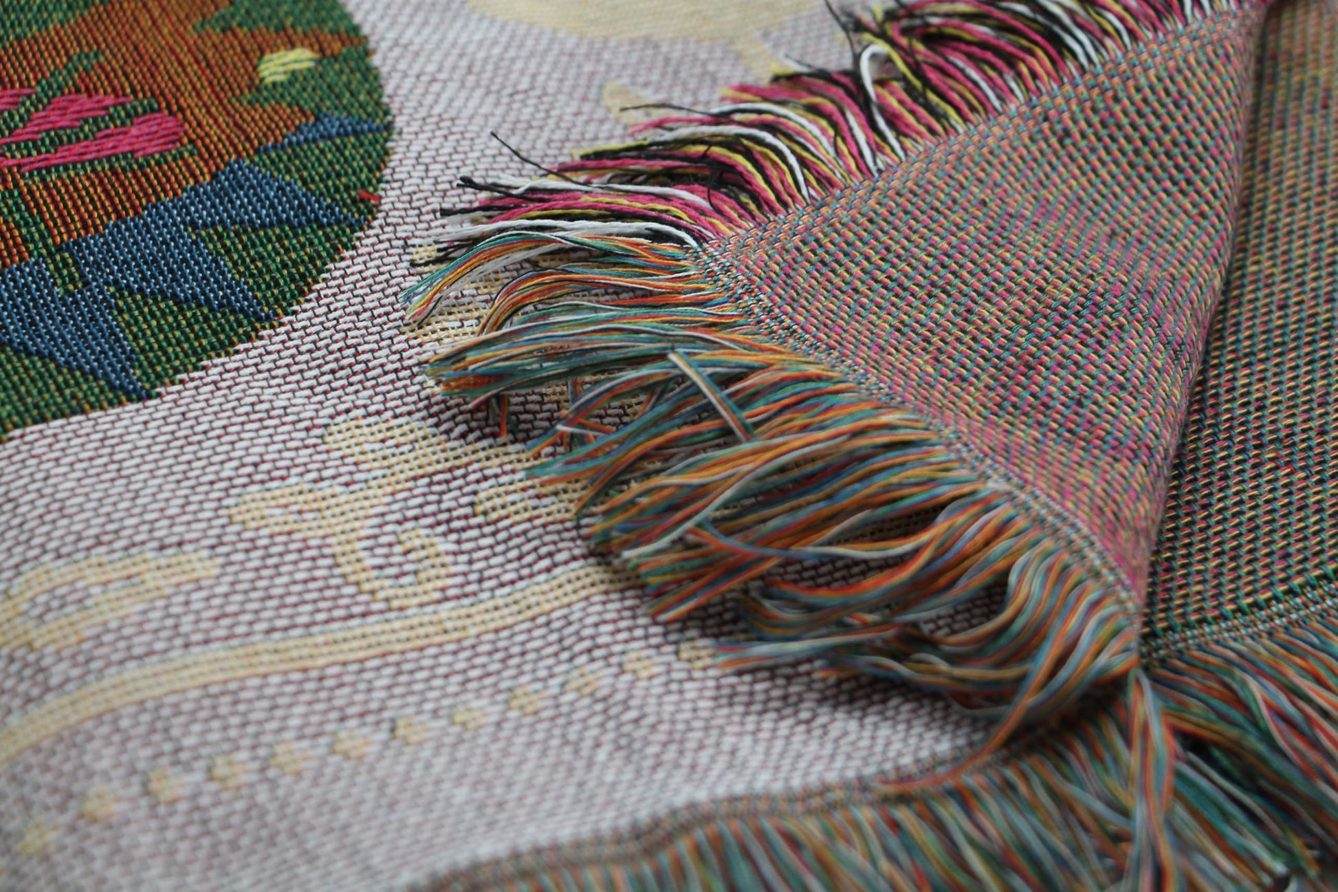 Close-up of the DETAIL Cat Oax Throwket by DETAIL, a woven textile inspired by Oaxacan throw blankets, featuring colorful fringed edges and intricate geometric and curved patterns. The fabric corner is folded over.
