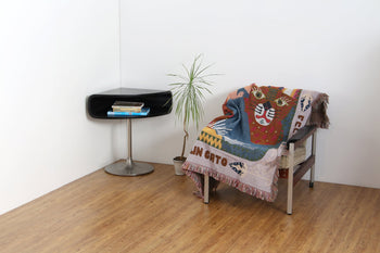 A modern, minimal room with wood flooring features a black side table with books, a potted plant, and a metal-framed chair draped with the DETAIL Cat Oax Throwket by DETAIL, a colorful Oaxacan blanket adorned with playful cats.