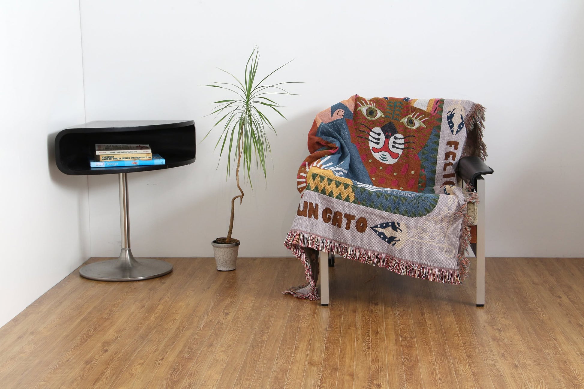 A modern minimalist room with a small potted plant, a black side table stacked with books, and a chair draped in the DETAIL Cat Oax Throwket by DETAIL, all set on wooden floors against crisp white walls.