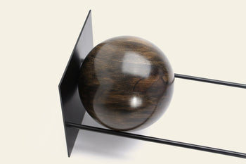 The DETAIL Slope Bookend by DETAIL features a polished wooden sphere against a vertical black metal plate, supported by two sleek parallel rods—a stylish minimalist accent on an off-white background.