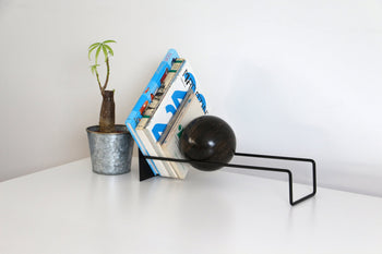 The DETAIL Slope Bookend by DETAIL holds several books alongside a small potted plant on a white surface, set against a plain white background.