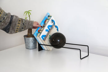 A hand arranges books on the DETAIL Slope Bookend by DETAIL, featuring a black metal frame and spherical accent, beside a small potted plant on a white surface against a plain wall.
