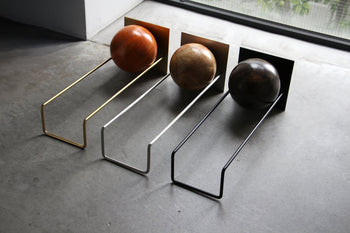 Three DETAIL Slope Bookends by DETAIL—gold, silver, and black metal stands—each display a large wooden sphere. Placed side by side on a concrete floor near a window, they elegantly match the varied wood tones.
