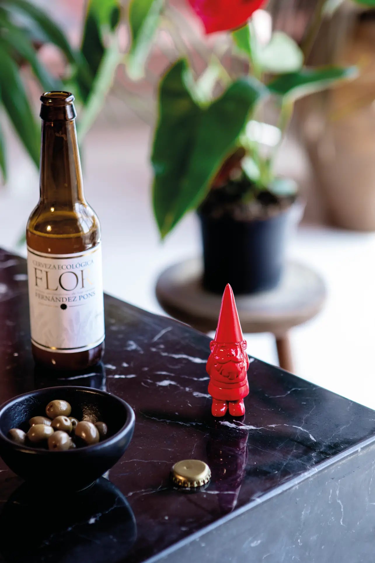 A brown glass beer bottle, a small black bowl of green olives, and the DOIY Gnome Bottle Opener by DOIY are arranged on a black marble table, with a gold bottle cap and a potted plant visible in the background.