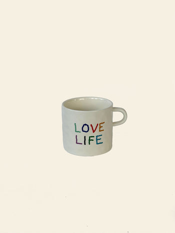 The David Hockney Love Life Mug by David Hockney is a white ceramic mug featuring the words “LOVE LIFE” in rainbow-colored letters, displayed against a plain, light beige background.