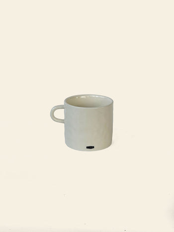 A white ceramic David Hockney Love Life Mug with a handle rests on a plain light background. A small black logo or text, characteristic of the David Hockney brand, appears near the base on the front side, lending an artistic touch to this classic ceramic piece.