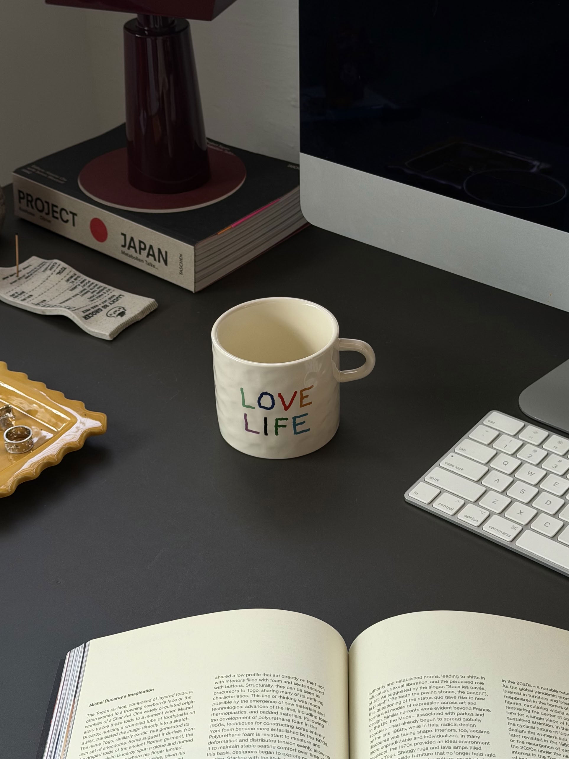 A David Hockney Love Life Mug by David Hockney sits on a dark desk next to a keyboard, an open book, a lamp, books, and a tray with jewelry.