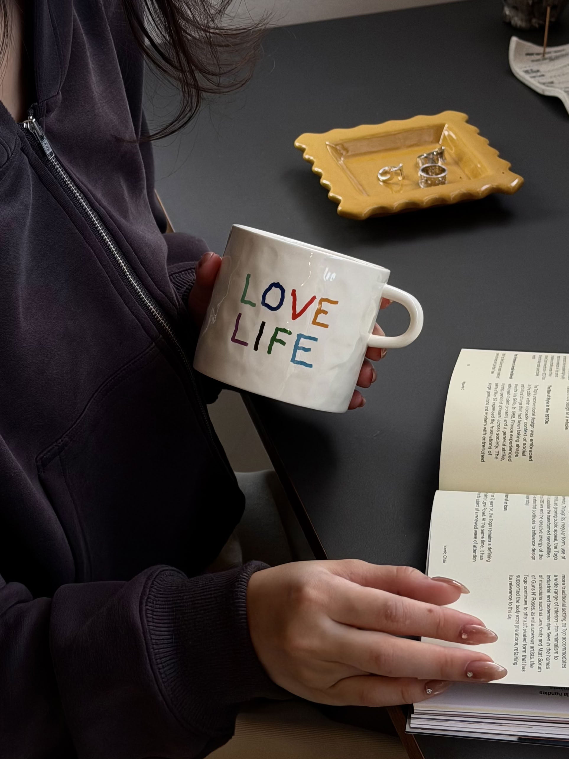 A person in a dark hoodie holds the David Hockney Love Life Mug by David Hockney and reads an open book at a table with a yellow dish holding rings.