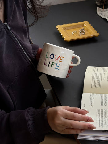 A person in a dark hoodie holds the David Hockney Love Life Mug by David Hockney and reads an open book at a table with a yellow dish holding rings.