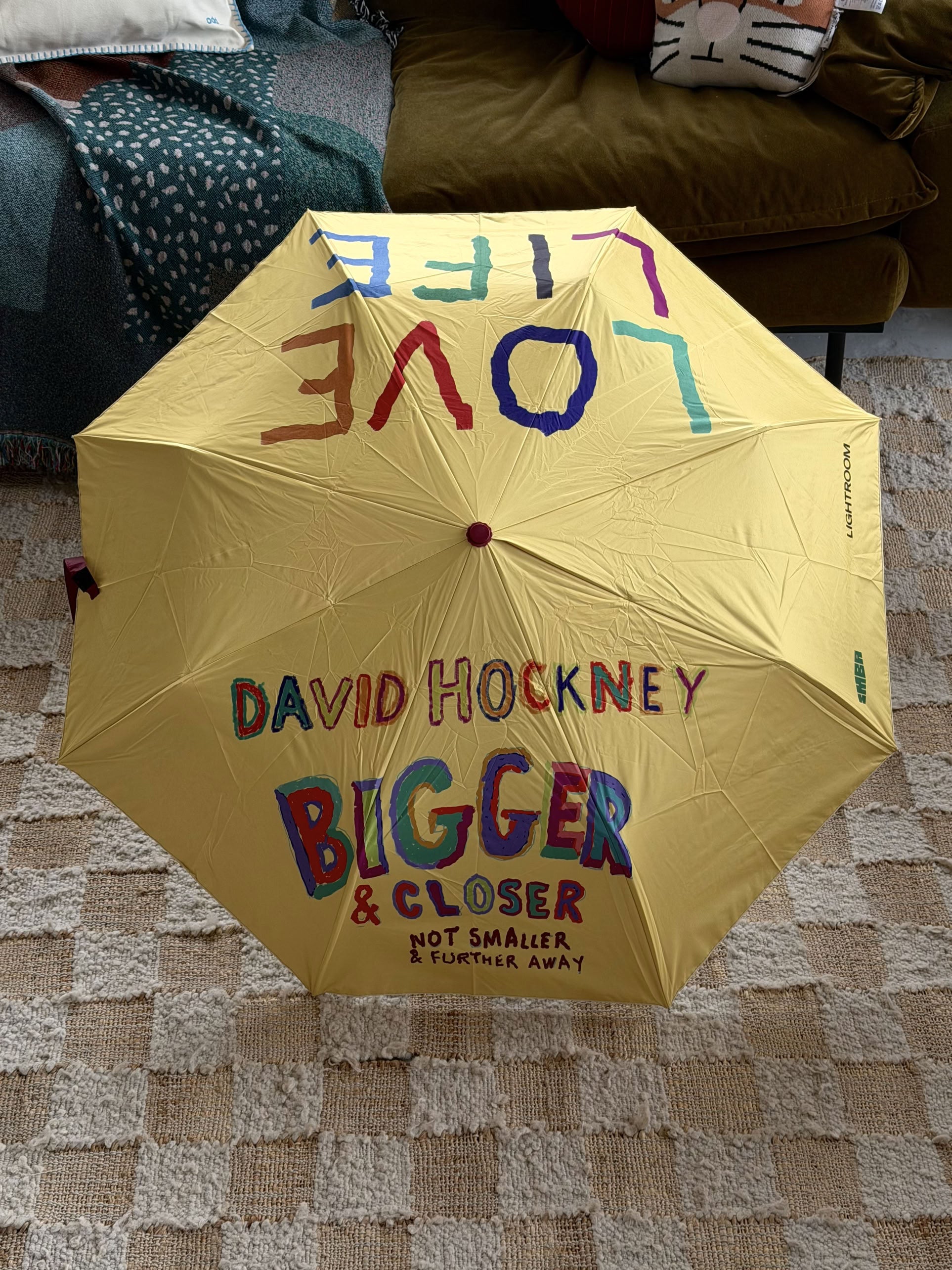 A yellow David Hockney Bigger & Closer Umbrella rests on a beige rug, featuring the phrases LOVE LIFE and DAVID HOCKNEY BIGGER & CLOSER NOT SMALLER & FURTHER AWAY in vibrant, playful lettering. In the background, a green couch and blue blanket highlight this cheerful David Hockney-branded umbrella as the focal point of the scene.