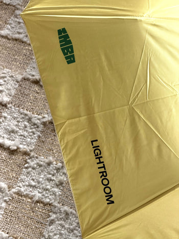 A David Hockney Bigger & Closer Umbrella by David Hockney, featuring a yellow canopy with “LIGHTROOM” and a reversed green “THEBA” printed on it in a style reminiscent of Hockney’s art, lies on a textured beige and white rug.
