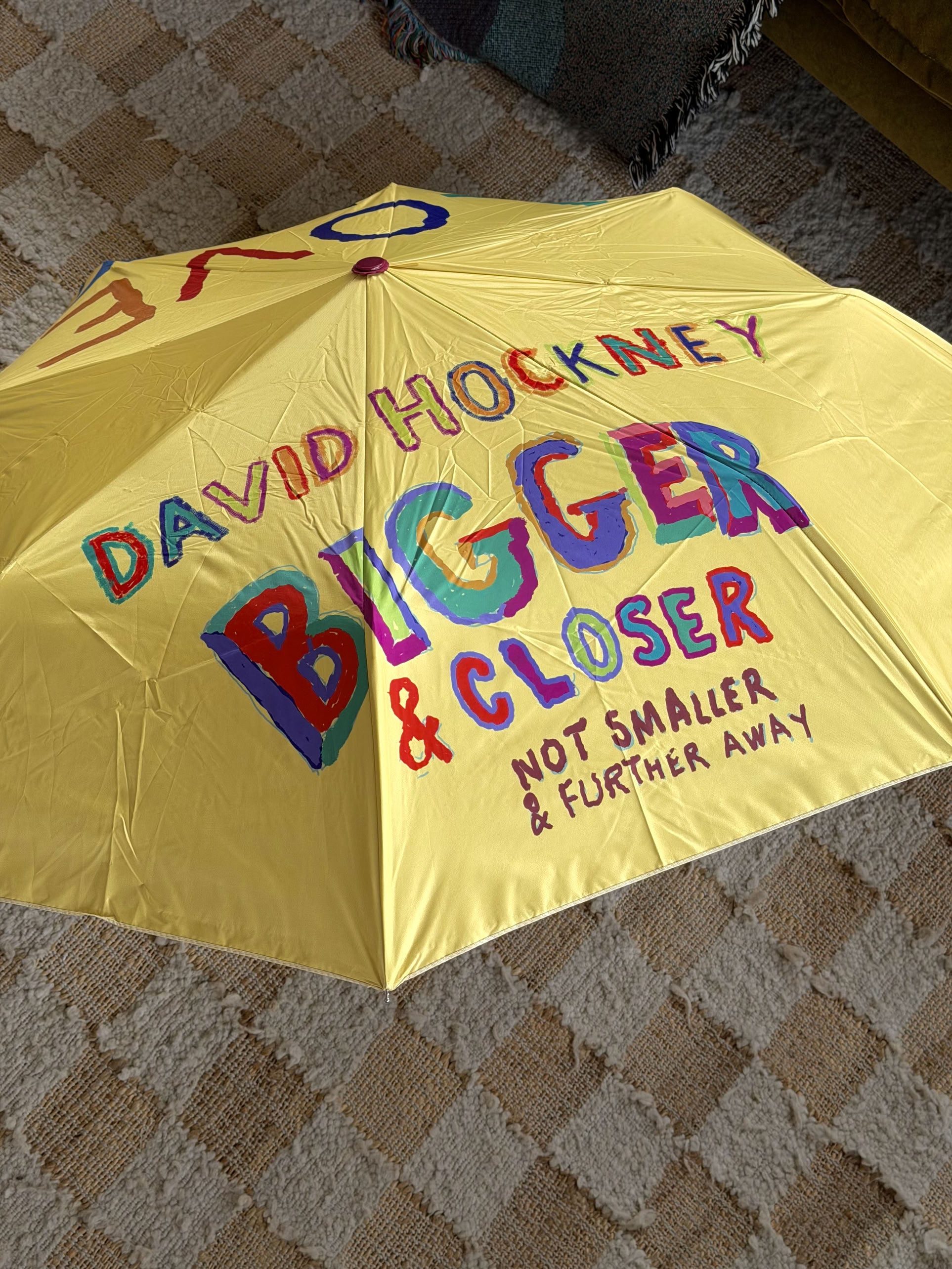 A yellow David Hockney Bigger & Closer Umbrella rests on a beige patterned carpet, its canopy featuring vibrant text that reads, “DAVID HOCKNEY BIGGER & CLOSER NOT SMALLER & FURTHER AWAY”—a bold accessory from the David Hockney brand guaranteed to brighten any space.