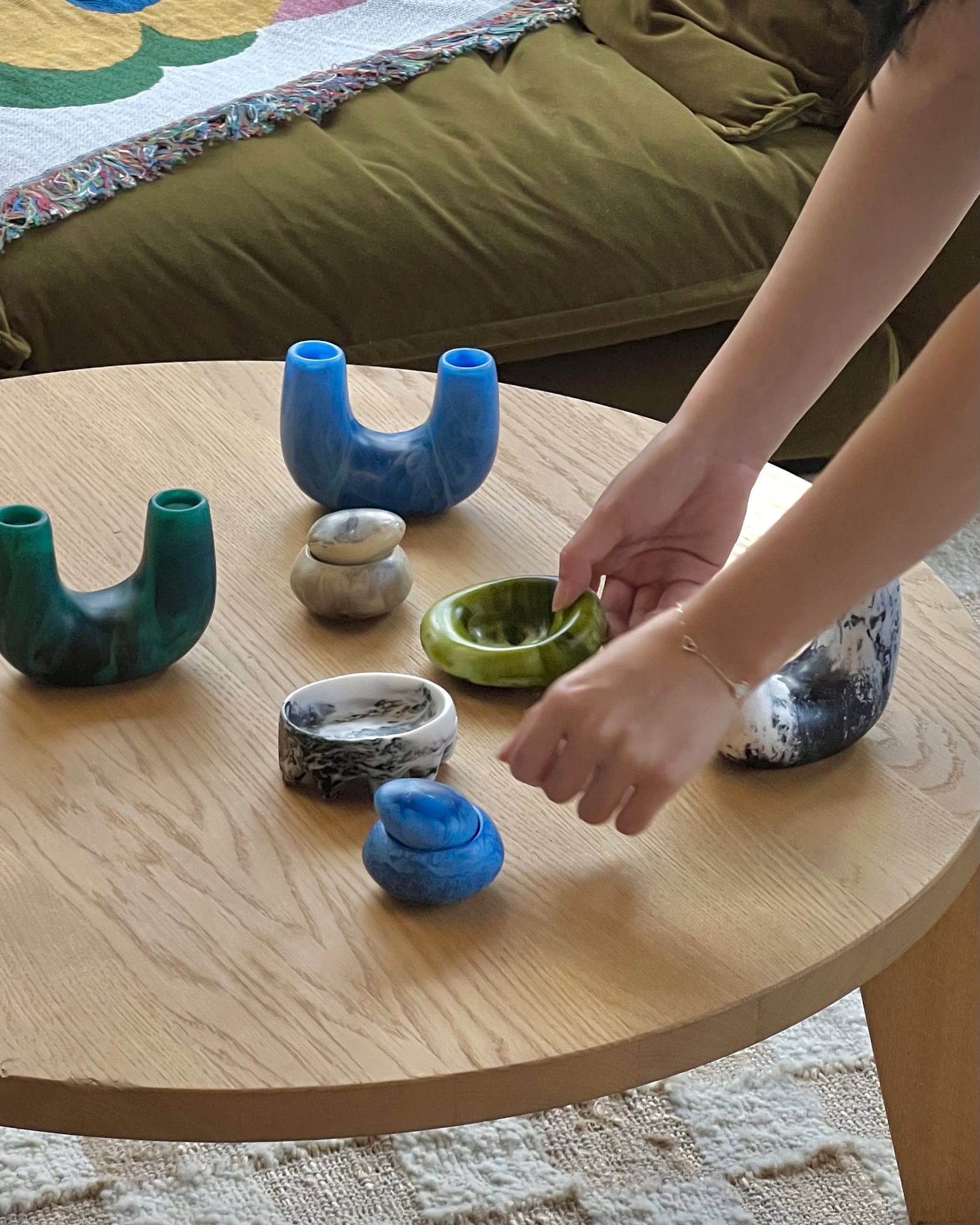 A person arranges colorful ceramic and stone objects, including the Dinosaur Designs Mini Branch Vase, on a round wooden coffee table. In the background sits a green couch with a patterned blanket.