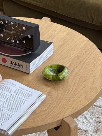 A wooden table displays an open book, a book titled JAPAN, a black digital clock, and a handcrafted Dinosaur Designs Small Drift Dish in green and white. A green couch by Dinosaur Designs enhances the organic feel in the background.