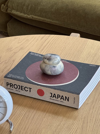 The Dinosaur Designs Small Pebble Jar, handmade from stone or ceramic with a sculptural design and matching lid, sits on a plain light beige background.